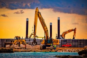 Two backhoes and one front loader working next to some water with a red hatchback car in the background