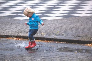 Blond child jumping in a puddle wearing jeans, a blue jacket, and red boots