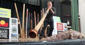 Man playing a digeridoo on stage with a fake alligator in front of him and posters behind him