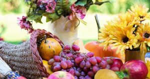 Cornocopia basket with gourds, apples, and grapes in it and a pitcher of yellow flowers next to it