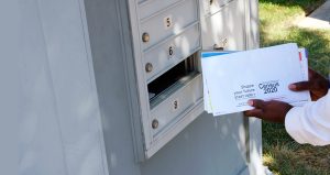 Two dark colored hands holding census 2020 envelopes in them in front of an open mailbox
