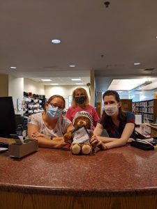 Three female librarians with a brown teddy bear dressed as a knight holding a sword and a sign that says Sir Nevins