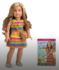 A blonde girl doll in a tropical pink, yellow and blue striped dress stands next to a book cover with the same girl, but human, standing in the same dress
