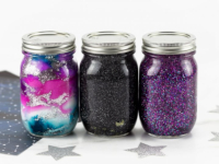 Three jars on a table, one filled with a purple sparkly material, one with a black sparkly material, and one with a pink white and turquoise sparkly material