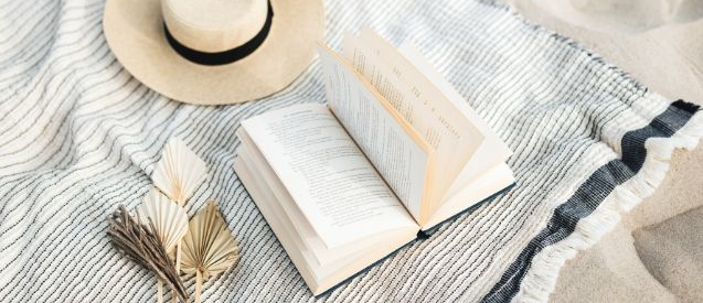 A white blanket with blue border that has a white sun hat a book flipping open and a grey/brown flower like thing laying on top of it.