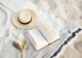 A white blanket with blue border that has a white sun hat a book flipping open and a grey/brown flower like thing laying on top of it.