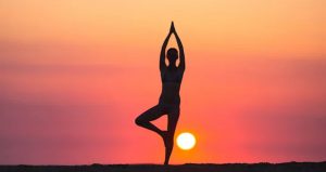 Silhouette of woman performing the one-legged prayer yoga pose in front of an orange sunset and the sun low in the sky