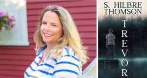 A Blonde woman standing in front of a red shingled side of a building next to a picture of her book "Trevor" by S. Hilbre Thomson