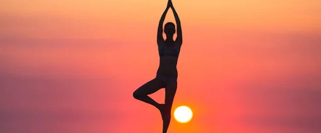Silhouette of woman performing the one-legged prayer yoga pose in front of an orange sunset and the sun low in the sky