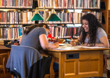 A picture of two girls writing at a desk with a bookshelf of books behind them and a old style green lamp next to them