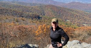 Woman in hat, black shirt, and khaki pants standing on a rock in front of green and yellow mountains