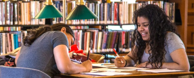 A picture of two girls writing at a desk with a bookshelf of books behind them and a old style green lamp next to them