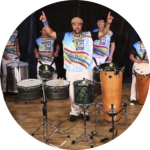 A man in a brightly colored shirt with his hands in his arms in the air and drums in front of him and his band