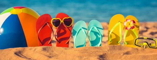 A beach ball, pair of red flip flops with sunglasses, pair of turquoise flip flops, pair of yellow flip flops, and a scuba face mask all stuck in the sand.
