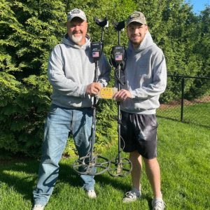 Two light skinned men in grey hoodies holding metal detectors in front of trees