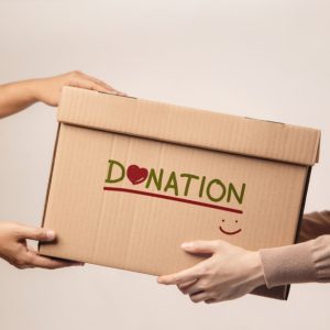 Light skinned arms handing a brown box with the word Donation on it and a smiley face to another person with light skinned hands
