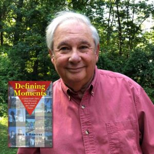 Hispanic looking white haired man in a pink collared shirt with the book 'Defining Moments' next to him