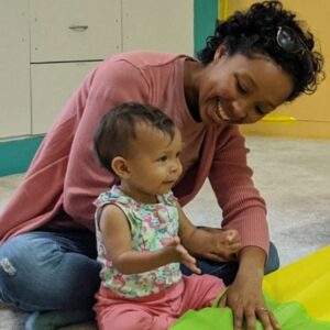 Dark skinned woman smiling down at a young child as they both sit on a colorful carpet