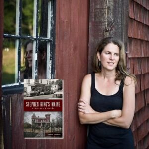 Light skinned woman in a black tank top standing in front of an old red house and door