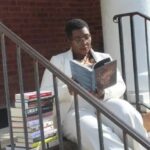 Black Experience of Revolution 654 Dark skinned woman in glasses and a white pant suit reading a book with a pile of books next to her