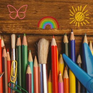 Colored pencils and paintbrush and scissors on a table with drawings of a butterfly rainbow and sun above them