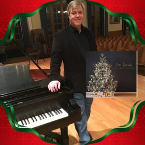 Light skinned man with blond grey hair in black shirt and jeans standing next to a piano on a stage