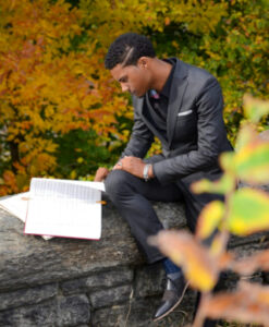 Black man sitting on a rock in a suit and reading a book of music