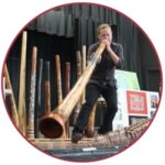 Light skinned man in dark clothes playing a digeridoo