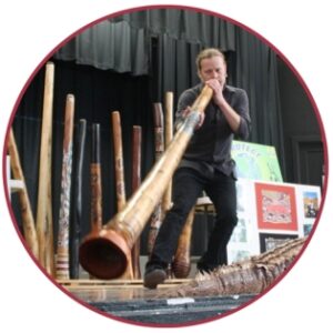 Light skinned man in dark clothes playing a digeridoo