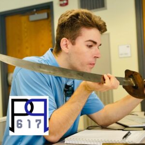 Young light skinned man with short brown hair holding, and staring intently at an old sword