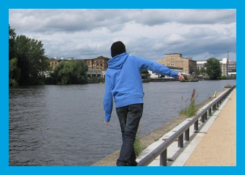Teen in a blue hoodie and black cap balancing on a railing next to a body of water