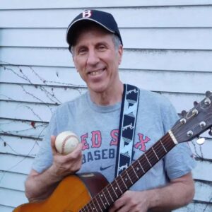 Light skinned older man wearing a Red Sox shirt and hat and holding a baseball and guitar