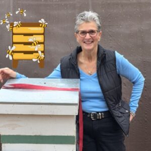 Light skinned woman with gray hair and glasses standing next to a bee hive