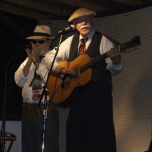 Two older men standing on stage, one with a guitar, the other with a harmonica