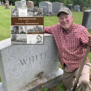 Light skin man in a red shirt kneeling next to a gravestone with the name White on it