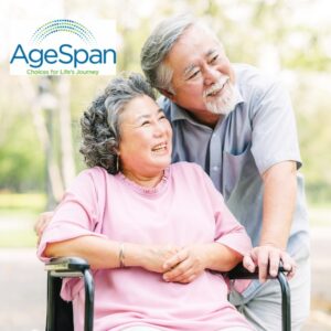 Older light skinned man standing next to older light skinned woman in wheelchair